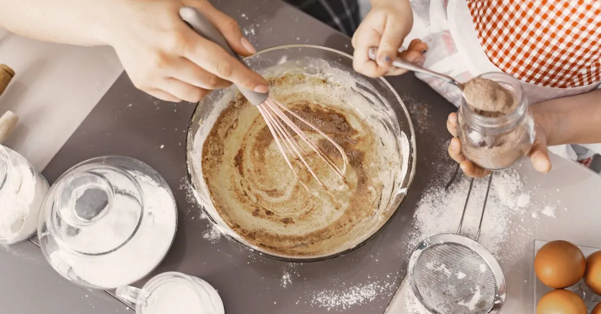 A close-up photo of a woman and child mixing ingredients in a bowl to make a cake. The woman is holding a whisk in one hand and the child is adding flour to the bowl with the other hand.
