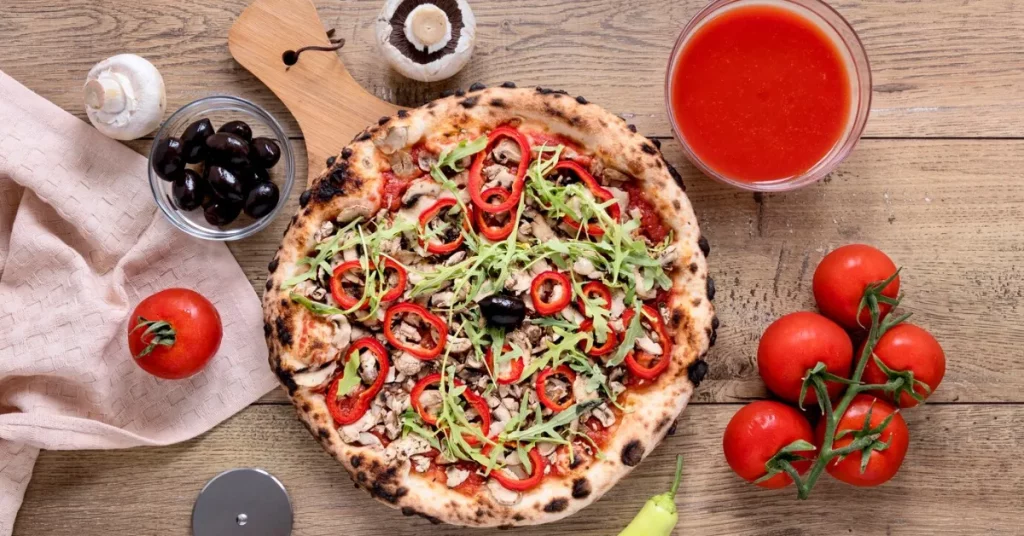 A Vegetarian Pizza on a wooden table surrounded by tomatoes,pepper, and a small bowl of olive.