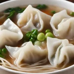 A close-up of a bowl of wonton soup with dumplings, chili sauce, and soy sauce.