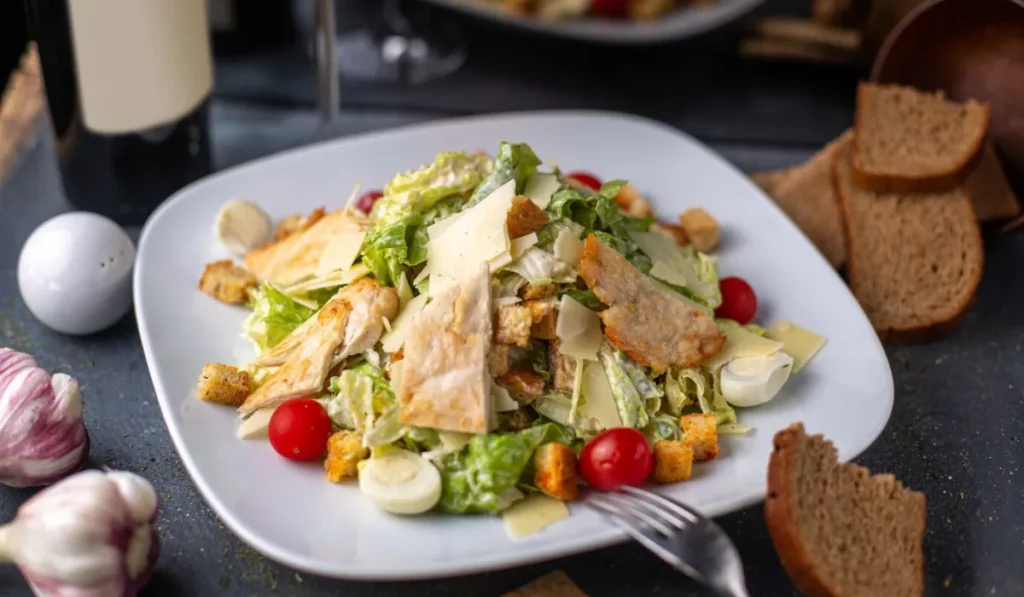 A close-up view of a plate of caesar salad with grilled chicken, parmesan cheese, croutons, and a light dressing. The salad is served on a white plate with a black rim. In the background, there is a blurred image of a restaurant setting.