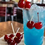 A refreshing blue cocktail, kenny chesney cooler recipe, in a tall glass with a straw and cherry on top, sitting on a wooden table
